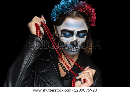 Creative makeup for Halloween party. Voodoo girl with picture makeup on her face, spooky scary skull. Holds beads jewelry. White teeth, black eyebrows drawn. Wreath of flowers, cool leather costume