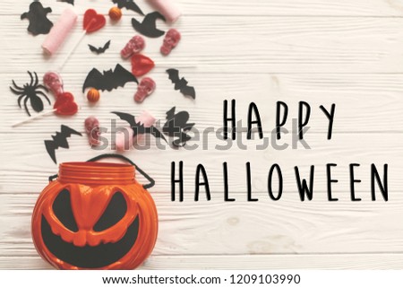 Happy Halloween text sign, flat lay. Jack o Lantern bucket with holiday candy, bats,spiders, skulls on white rustic wood. Space for text. Season's greeting card