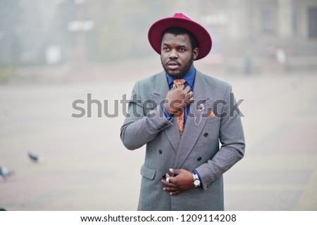 Stylish African American man model in gray jacket tie and red hat posed on street with fog.