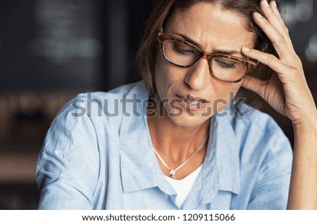 Portrait of stressed mature woman with hand on head looking down. Worried woman wearing spectacles. Tired lady having headache sitting indoors. Royalty-Free Stock Photo #1209115066