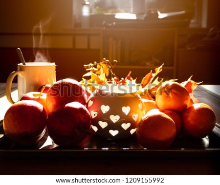 A fruit tray with apples and oranges, a cute candle holder, fall leaves, twigs of berries and a steaming mug of tea on the side. Warm sunlight streaming in from the window. A picture of autumn colors.