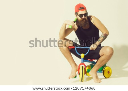 bearded man, long beard, brutal caucasian hipster with moustache in black vest, sunglasses, red cap holds green baseball bat with serious face sitting on colorfyl bicycle toy on grey background