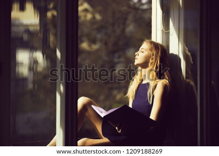 Daydreaming and relaxing. Pretty girl or young woman, student, teenager, with cute young face and blond, long hair in dress sleeping with book at open window on sill on sunny day on urban background