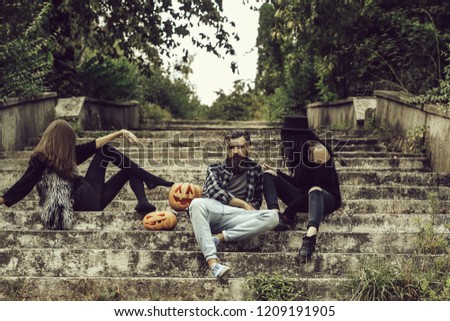 young halloween bearded man with beard and mustache and two girls in black witch hat tights and jeans sit on stony stairs with traditional autumn holiday symbol of orange spooky pumpkin outdoor.