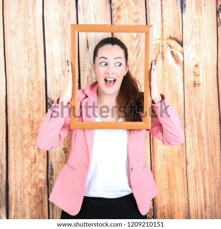 Excited young woman holding an empty picture frame while looking to one side against a wooden background