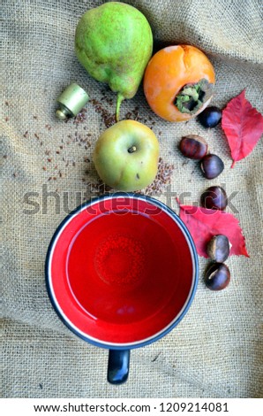 Autumn relaxation still life. Close up of cup of herbal tea (shabby red and black), persimmon, roasted chestnuts, green apple, pear, flaxseeds, red leaves, balance weight, on jute background. Natural.