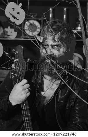 Demon with horns and evil face holds black electric guitar. Devil or monster playing instrument. Man wearing scary makeup behind Halloween pumpkins and bats decor. Halloween costume party concept