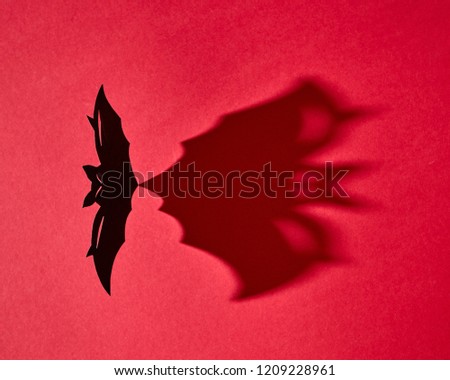 Black handmade craft paper bat with shadow on red background and copy space. Creative mystical halloween card. Flat lay