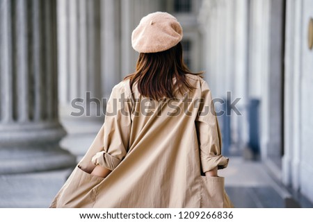 A shot of the back of a stylish Chinese Asian woman in an elegant French-inspired outfit (with a khaki trench coat and beret) in the corridor of a trendy building during the day. Royalty-Free Stock Photo #1209266836