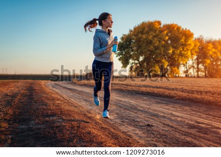 Woman running holding bottle of water in autumn field at sunset. Healthy lifestyle concept. Active sportive people Royalty-Free Stock Photo #1209273016