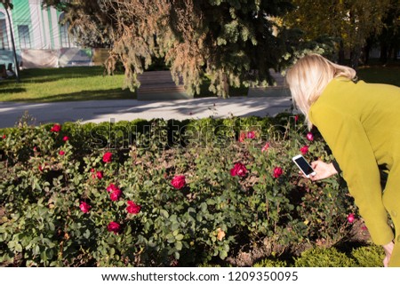 Young woman in mustard coat using smartphone to take a photo of beautiful flowers roses in autumn park