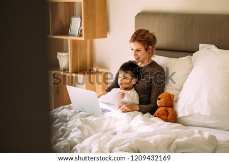 Young mother with her beloved son in the bedroom on the bed watching a movie on laptop. Adorable boy sitting on bed with mother and watching cartoons on laptop computer.