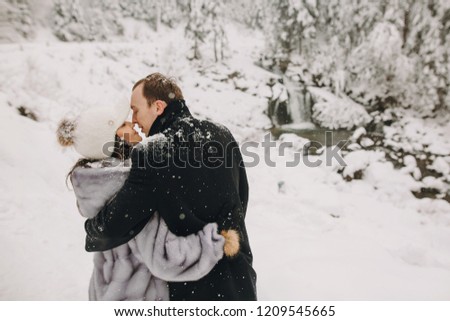 Stylish couple embracing in winter snowy mountains. Happy romantic man and woman in luxury clothes gently hugging at waterfall in snow. Holiday getaway together
