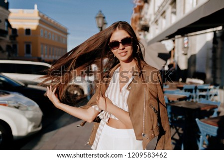 Pretty laughing woman with long hair has a good time in autumn weekend. Outdoor portrait of lovable trendy lady playing with her hair and has fun in sunlight on avenue