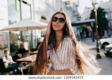 Amazing dark-haired lady in good mood wearing sunglasses and fashion t-shirt posing outside in sunset. Enthusiastic young woman chilling outdoor in weekend.