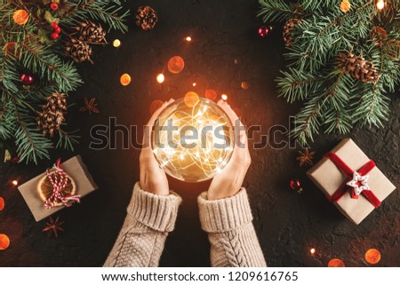 Female hands holding Christmas glowing ball on holiday background with Fir branches, gifts. Xmas and Happy New Year card, bokeh, sparking. Flat lay, top view
