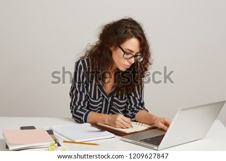 Closeup of a young woman makes notes and keeps one arm on a laptop keyboard, sitting at the table, looking thoughtful, wears white and black stripped shirt and glasses isolated over white background