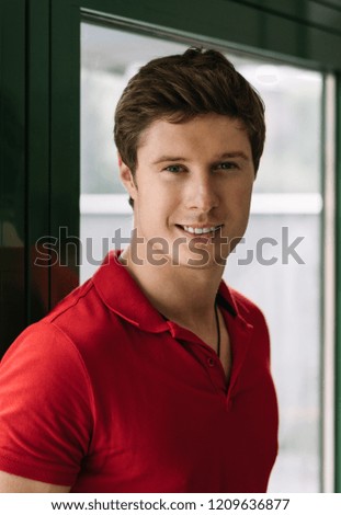 Young handsome guy wearing stylish casual red t shirt posing for pictures and smiling, standing in loft cafe near window. Portrait of happy successful man waiting taxi indoors. 