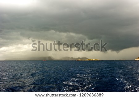 Picture taken from a yacht sailing away from the storm. 
Dark, heavy clouds hanging and rain pouring over an island in the distance; deep blue and stormy sea. 