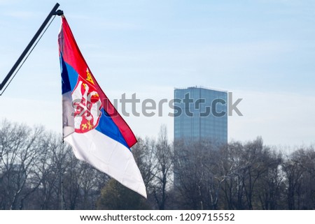 Photo of the official flag of the republic of Serbia, in front of a business building in Belgrade, Serbia, with a backround of blue sky and clouds

