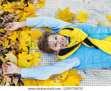 cute boy smiles in blue shirt and yellow scarf lays on beige plaid with maple yellow leaves. autumn mood beautiful day. Golden fall in still life. Bright Fall image.  