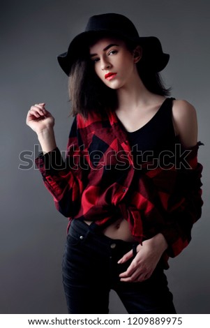 Beautiful young woman posing in red plaid shirt and black hat, isolated over grey background
