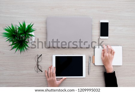 The woman is working with various office gadgets like labtop, tablet and smartphone. She is  writing on the small notebook  on the wooden desk in office.