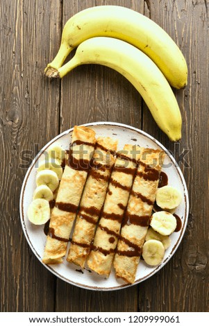 Tasty crepes roll with banana slices and chocolate syrup on wooden table. Top view, flat lay
