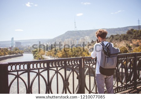 Young curly traveler taking photo with camera in Tbilisi, Georgia. City viewfrom bridge.