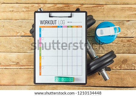 Healthy lifestyle concept. Mock up on workout and fitness dieting diary with copy space. Workout log sheet, blue shaker, measuring tape and dumbbells on a wooden background