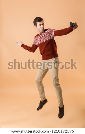 Full length portrait a handsome young man jumping isolated over beige background, taking selfie with mobile phone