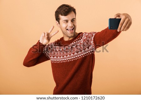 Portrait a smiling young man standing isolated over beige background, taking selfie with mobile phone