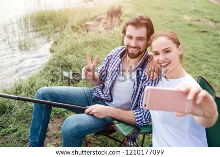 Another picture of girl taking selfie of herself and her husband. She is doing that with frontal camera on phone. Guy is smiling and showing the piece symbol. Also he is holding the end of fish-rod.