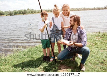 A picture of whole family standing on the grass and looking at fish that dad has caught. Kids and woman look amazed. They are happy. Man is lookiing at them and smiling.
