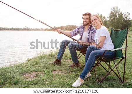 A picture of awesome young people sitting together at river shore and smiling on camera. Guy is holding huge and long fish-rod while his wife is leaning her head on his shoulder.