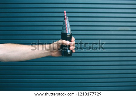 A picture of man's hand holding small bottle of coca-cola with straw in it. Isolated on striped and blue background.