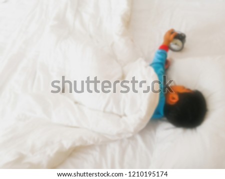 This image is deisgn to blur.A boy wearing a blue t-shirt lies under the blanket. Hand holding a black watch.Children sleep on a white bed.This picture was shoot from top view.