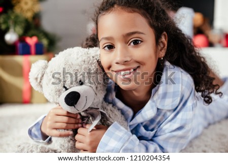 portrait of adorable happy african american child in pajamas with teddy bear looking at camera at home