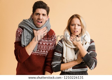 Portrait of a frozen young couple dressed in sweaters and scarves standing together isolated over beige background, shaking