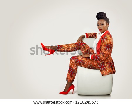 Confident African business woman  wearing an African print suit and heels with a sophisticated hair style sitting in a clean space with her leg propped up. Royalty-Free Stock Photo #1210245673