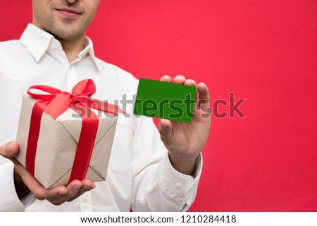 Attractive businessman in white shirt showing business card to the camera, holding a box with a gift with a red ribbon, bright red background, with copy space, for advertisement, front view