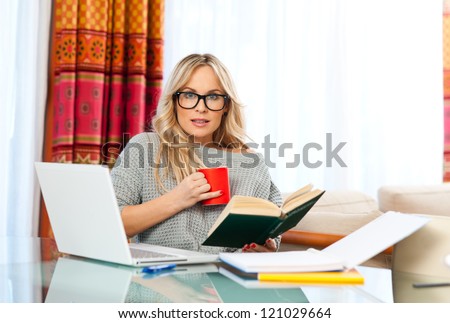 attractive woman with cup of coffee, book and laptop sitting at her home desk and working