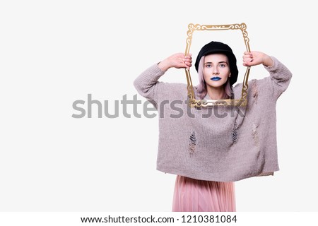 Portrait of young emotional attractive woman in black cap, torn sweater, with blue lips and pink hair, she is holding imitation of picture frame, isolated on white background.