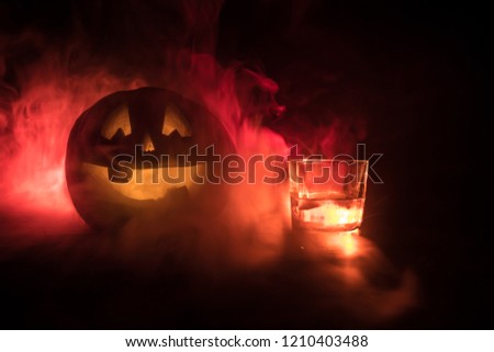 Halloween pumpkin with carved face and glass of whiskey with ice on a dark toned foggy background. Decorated. Selective focus