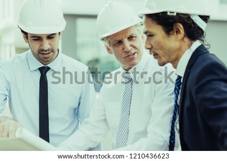 Closeup portrait of three serious diverse business people wearing helmets, holding blueprint, discussing it and standing outdoors. Construction project concept.