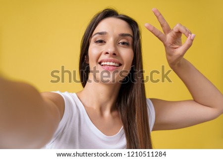 Pleasant attractive girl making selfie in studio and laughing. Good-looking young woman with brown hair taking picture of herself on bright yellow background.