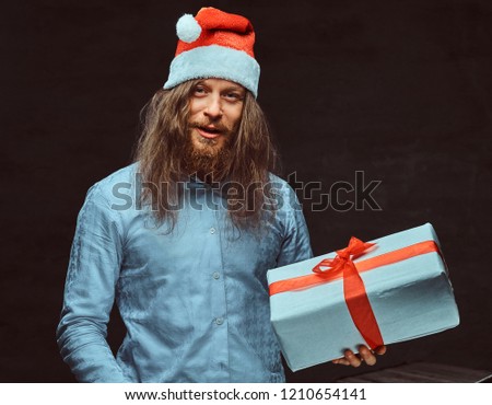 Happy male with long hair and beard in blue shirt and red Santa hat holds gift box.
