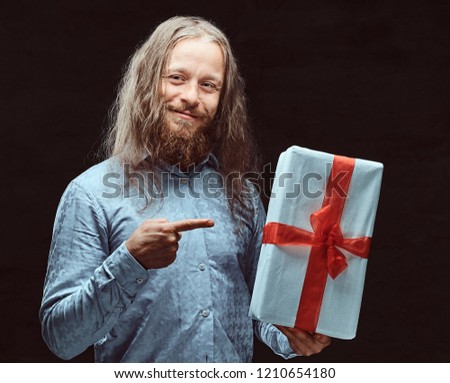 Happy male with long hair and beard in blue shirt pointing on gift box in his hand. Christmas concept.