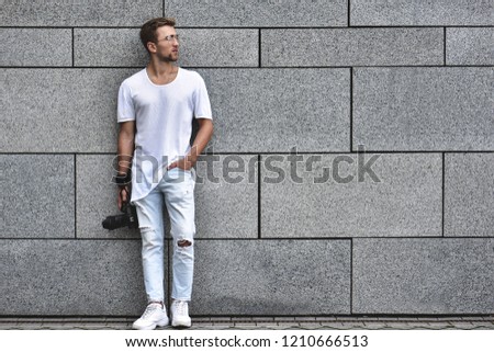 Picture of handsome Caucasian guy on grey textured wall in white T-shirt and white jeans with camera hanging on strap on his arm.