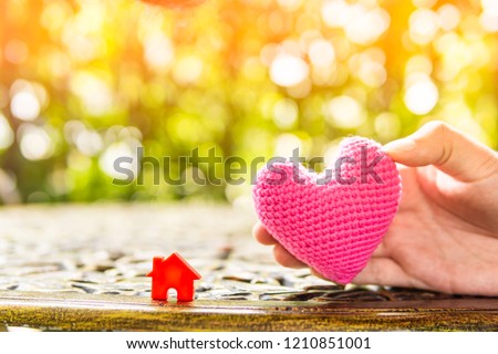 Hand are hold pink heart and home model.Give love to the family for happiness..Have background in garden and light of sun.